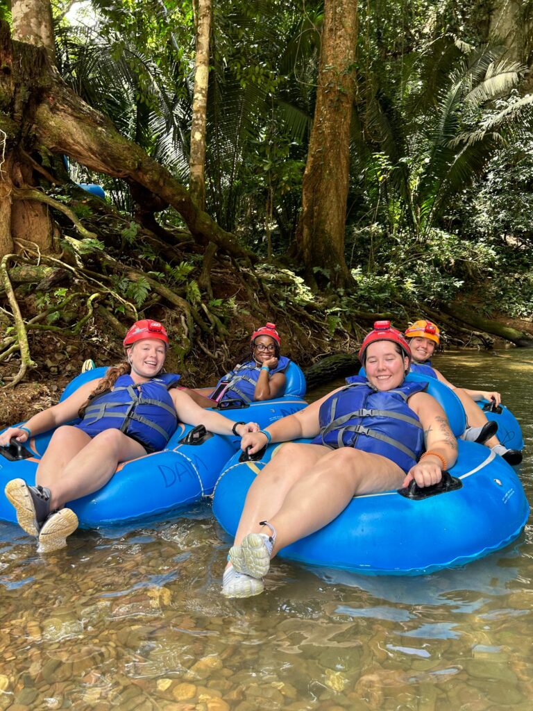 Participants float on river tubes beside a wooded bank.