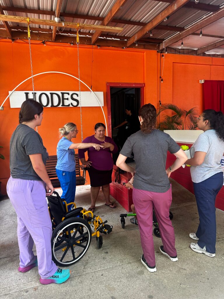 A group practices a supported standing activity beside a wheelchair.
