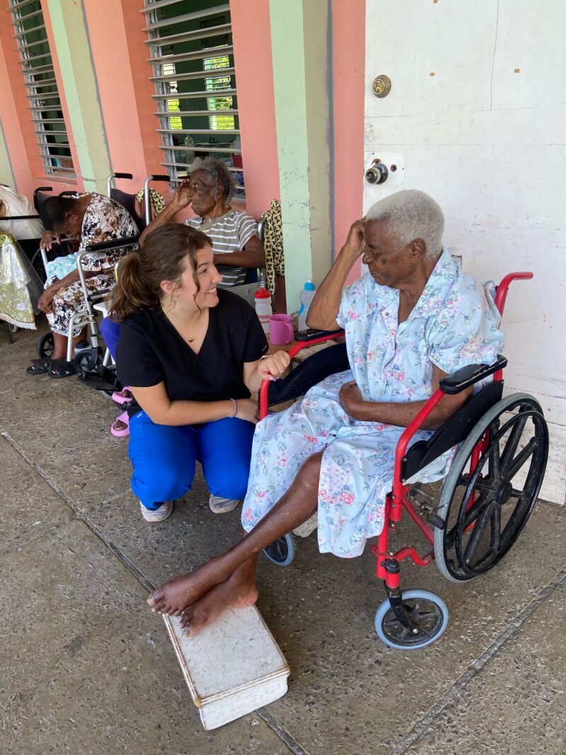 A young female therapy student in blue scrubs kneels to hold the hand of an elderly woman in a red wheelchair at an outdoor healthcare facility. The student is smiling and engaging in supportive patient care, while other elderly residents in wheelchairs are visible in the background against a pink and green building.